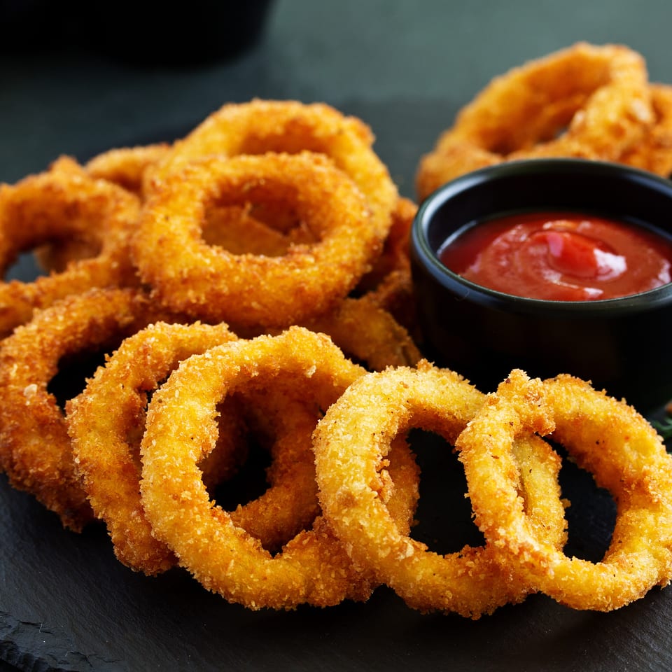 Crispy golden onion rings piled on a dark slate board with a small bowl of ketchup.