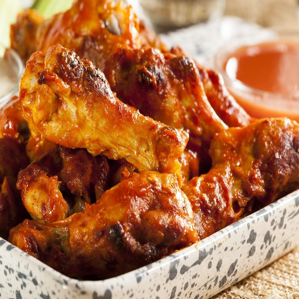 Spicy buffalo chicken wings in a speckled white bowl, with ranch and hot sauce in the background.