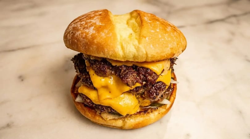 A large double cheeseburger with melted cheese, two patties, and a soft bun on a marble surface.